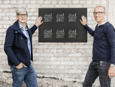 Fotograf Alexander Feig und Jürgen Frischmann stehen vor Backsteinwand Feigefotodesign